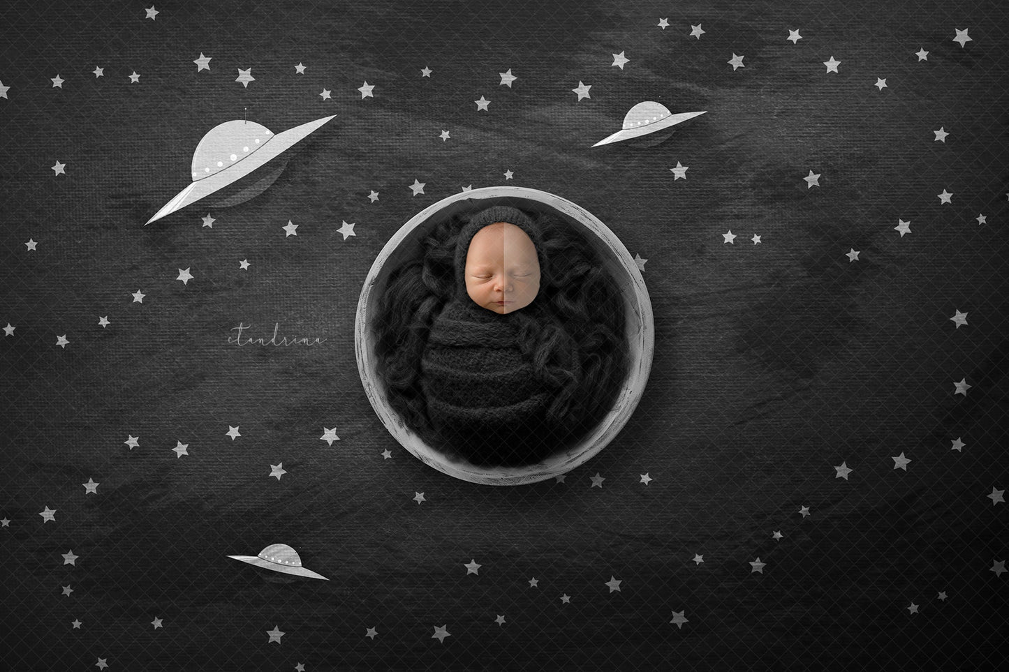 A newborn baby wrapped in a black fabric inside of a distressed white painted wooden bowl on a black brushed background that has pictures of little ufos and many stars